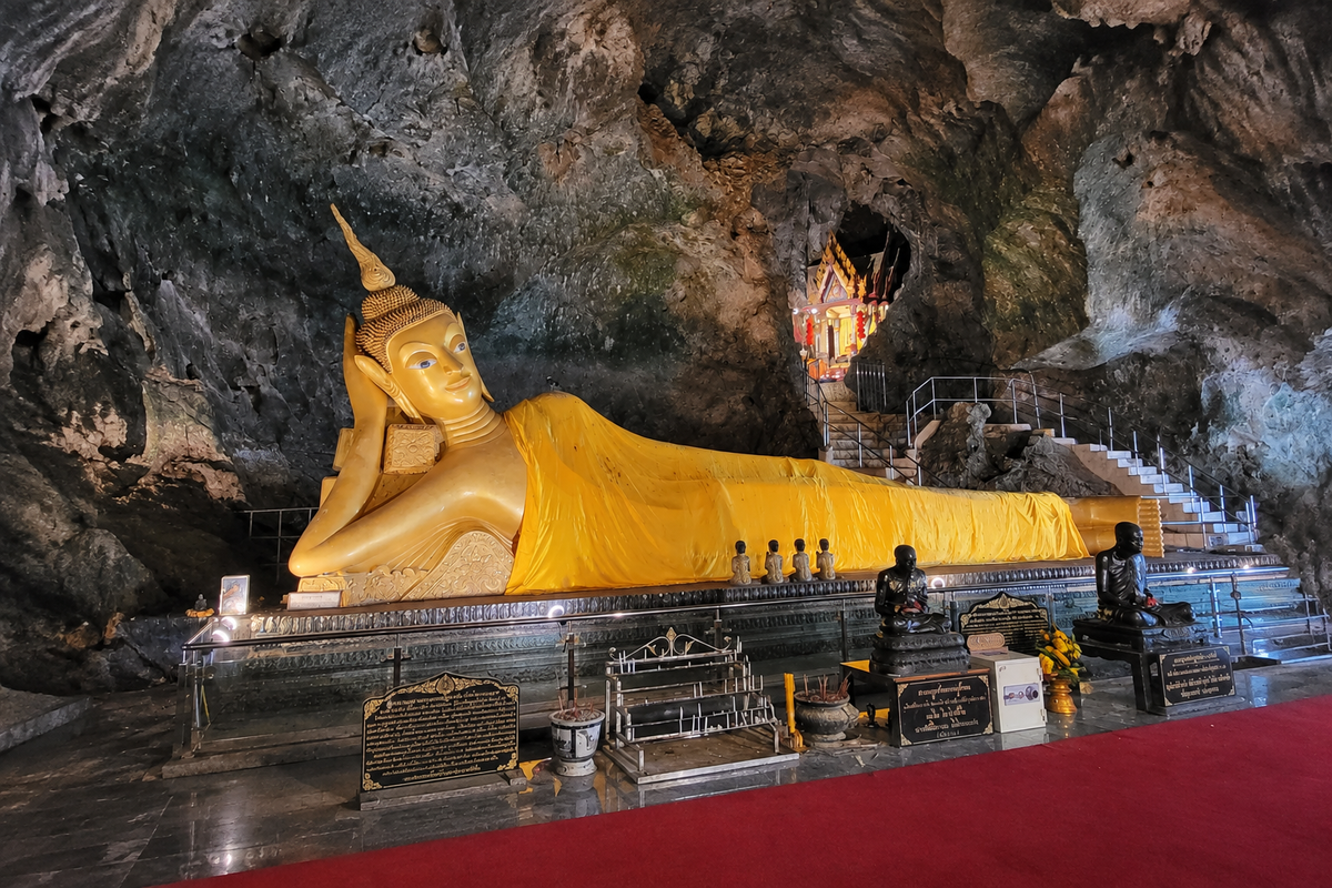 Liegender, goldener Buddha in einer natürlichen Höhle in Thailand mit kleinen Buddhafiguren und einem Altar im Vordergrund.